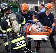 Firefighters rescue a woman, 75, from a house that exploded in flames yesterday. A neighbor kicked in the windows to help.