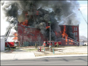 Quint 5 is positioned at the east (B) side of the structure. The elevated aerial stream and hand lines from this vehicle were alternately played upon the exposure and the fire building.