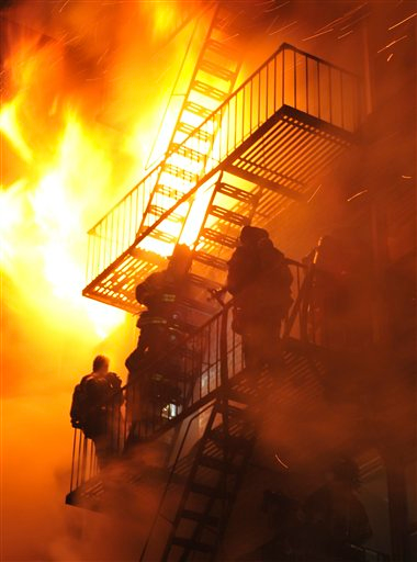 FDNY Firefighters stand on the fire escape as winds whip the flames from a five-alarm fire in Brooklyn.