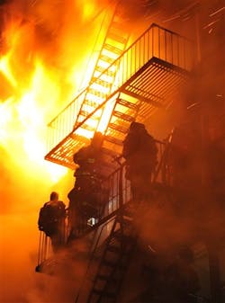 FDNY Firefighters stand on the fire escape as winds whip the flames from a five-alarm fire in Brooklyn. FDNY Firefighters stand on the fire escape as winds whip the flames from a five-alarm fire in Brooklyn.