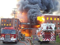 The fire is quickly jumping to an adjoining factory to the right. A mutual aid company is setting up defensive ladder operations, supported by Salamanca&rsquo;s first-arriving engine.
