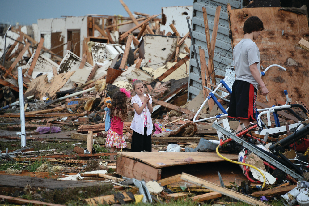 Children wait outside of what was once their home while their father searches in debris.
