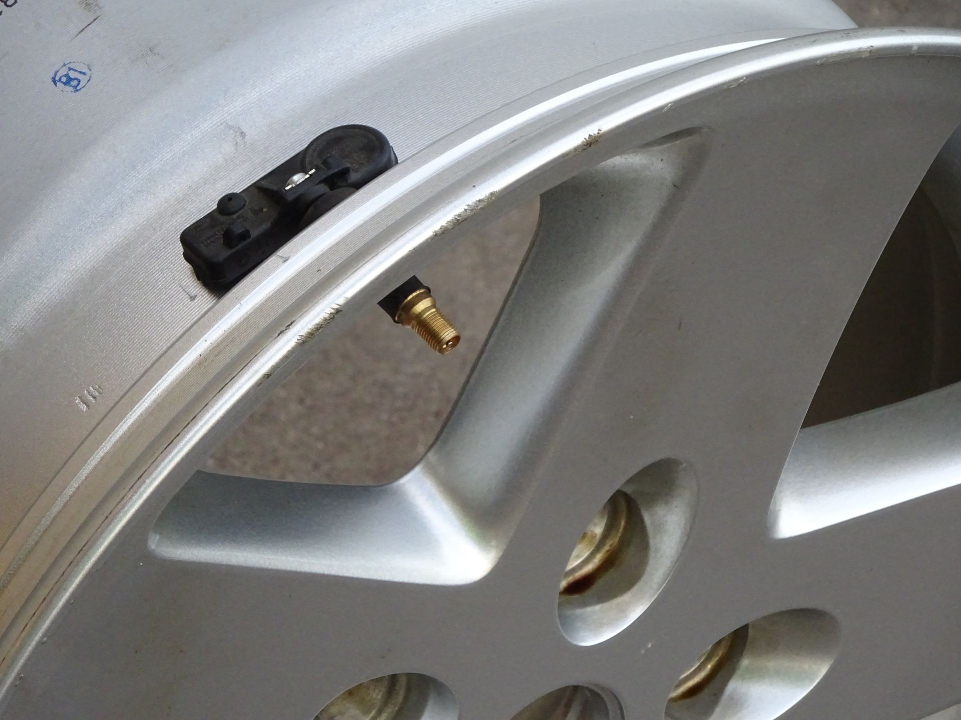 A tire-pressure monitoring unit is shown attached to a wheel (with the tire removed) to show the large transmitter unit inside the rim and the valve stem sticking outside of the wheel.