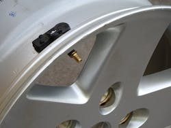 A tire-pressure monitoring unit is shown attached to a wheel (with the tire removed) to show the large transmitter unit inside the rim and the valve stem sticking outside of the wheel.