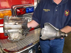 The large, stored gas inflator that ruptured and flew out of the windshield during the fire in the Dodge Neon is on the right. The passenger front-airbag mounting bracket is on the left. Both these pieces were found 120 feet ahead of the vehicle after the fire was extinguished.