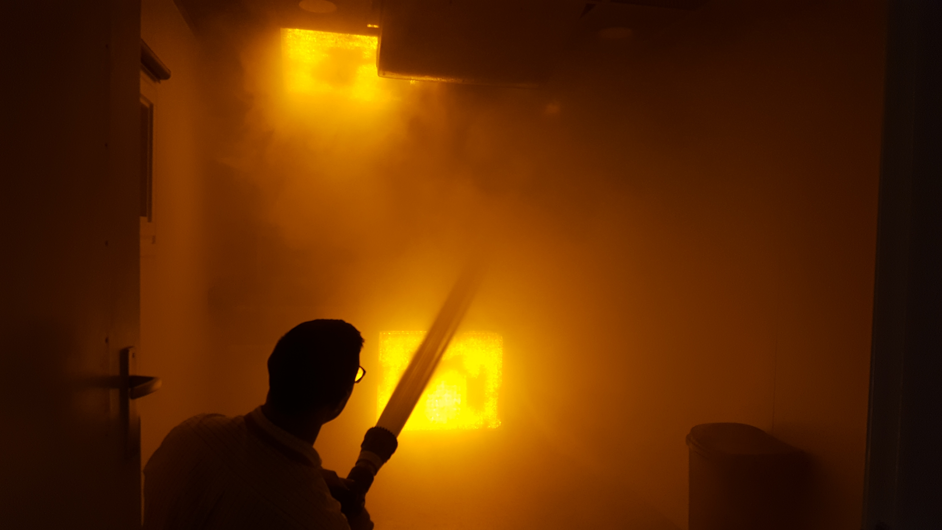 The man in the photo is not really in a fire building without turnout gear and SCBA. Rather, it's a simulated room and contents fire in BullEx's show booth and the man 'extinguishing' the fire is a representative of the company. BullEx's simulated fire board reacts to water streams, darkens down and eventually goes out with sufficient application of water. Smoke machines provide added scene realism.