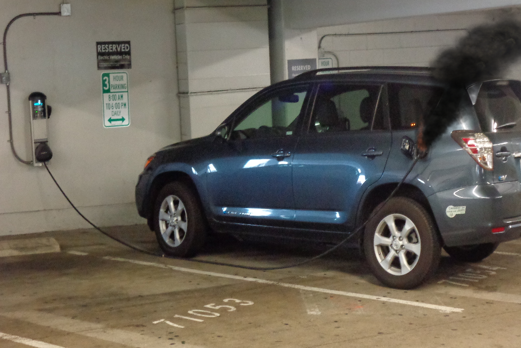 This electric plug-in vehicle is in a public parking garage. It is plugged into the wall-mounted charging station. A fire is evident at the end of the charging cable where it is connected to the charging receptacle on the vehicle. Recommended tactics include: Isolate and secure the immediate area, request power shutdown, protect exposures, chock wheels of vehicle if safe to do so, and initiate additional activities as appropriate once power is confirmed shut off to the charging station.