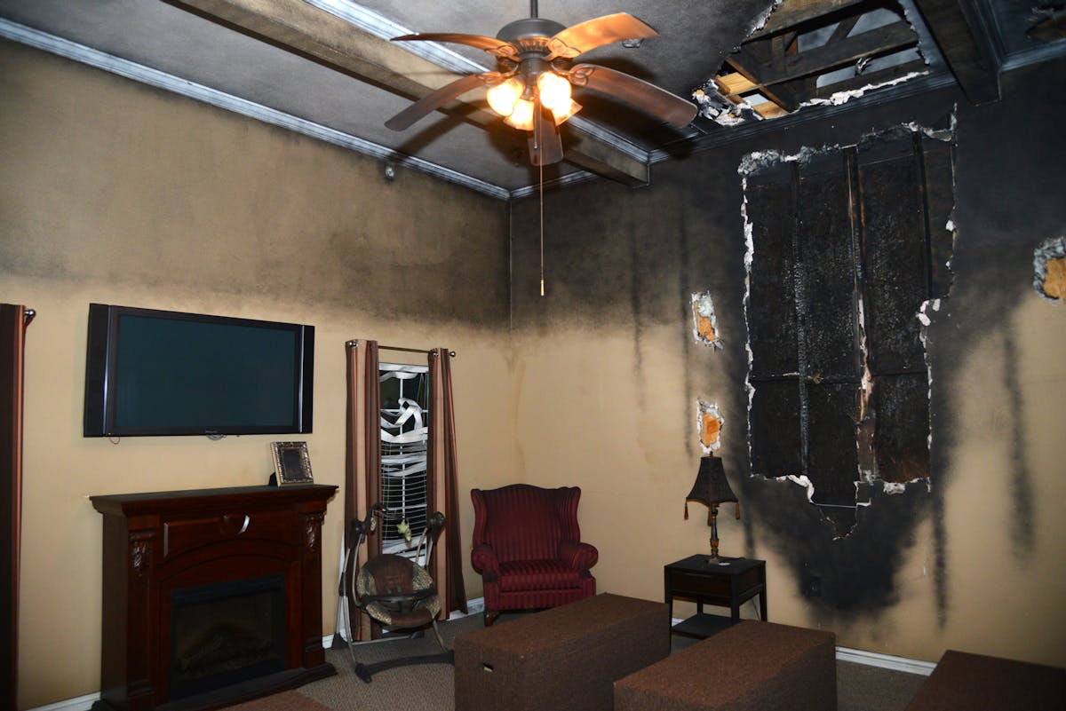 This high-tech living room features a ceiling and wall that have been scorched and ripped apart. The upper half of the room is covered in soot and items are melted, showing the effects of heat.