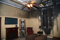 This high-tech living room features a ceiling and wall that have been scorched and ripped apart. The upper half of the room is covered in soot and items are melted, showing the effects of heat.