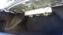 The two black trunk torsion rods of this KIA sedan are visible beneath the rear speaker deck. Note the mild steel mounting bracket for the torsion rods at the left of this image. This is where they can be pried free of the mount and removed.