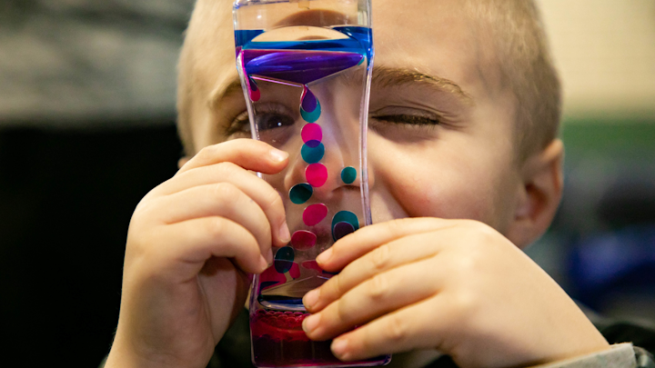 Carter Severs, 5, looks through a glass toy with pink and blue goo inside that floats around, similar to a lava lamp. The tool is part of new autism sensory kits, called Carter Kits after the boy.