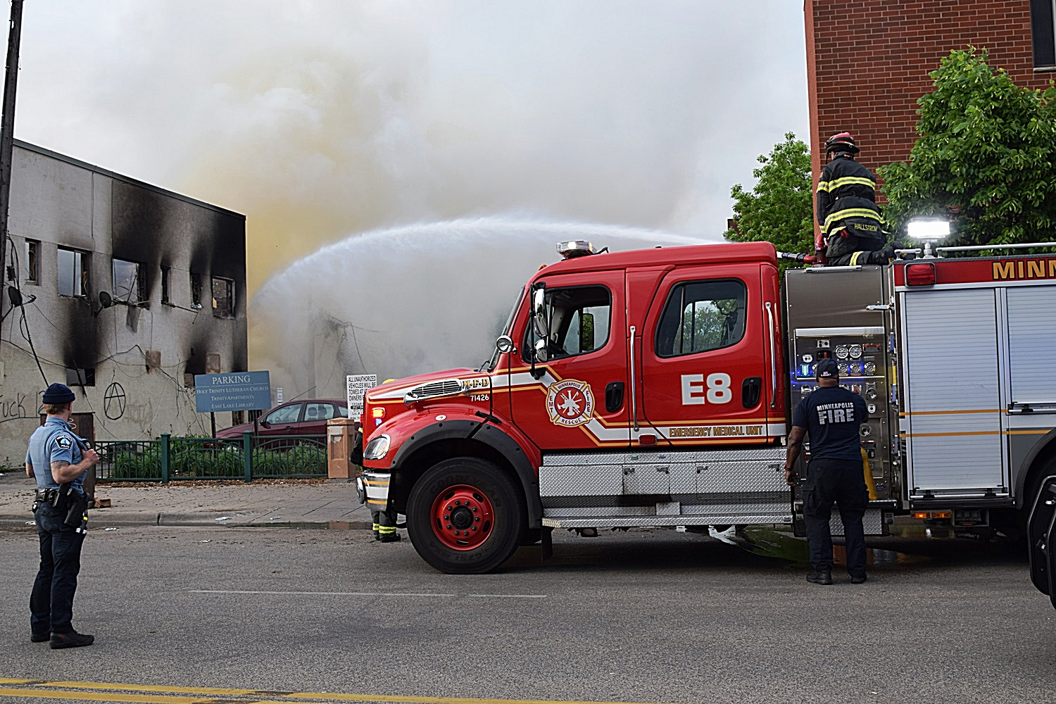 A Minneapolis police officer watches over Minneapolis firefighters who douse the aftermath of a fire following riots in May.