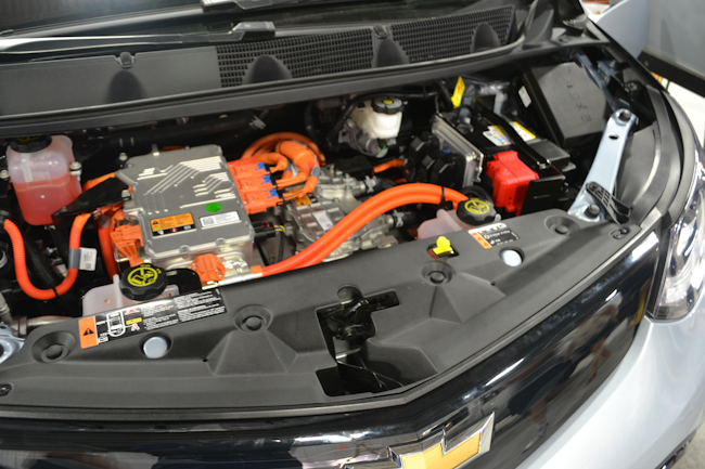 A 1st Responder Cut Label (yellow) for this Chevrolet Bolt EV is visible at upper right along the firewall. The larger label at lower left on the top of the radiator trim piece indicates that there is a 400-volt lithium-ion battery onboard and that the 12-volt battery is in this front compartment, somewhat buried on the lower left side.