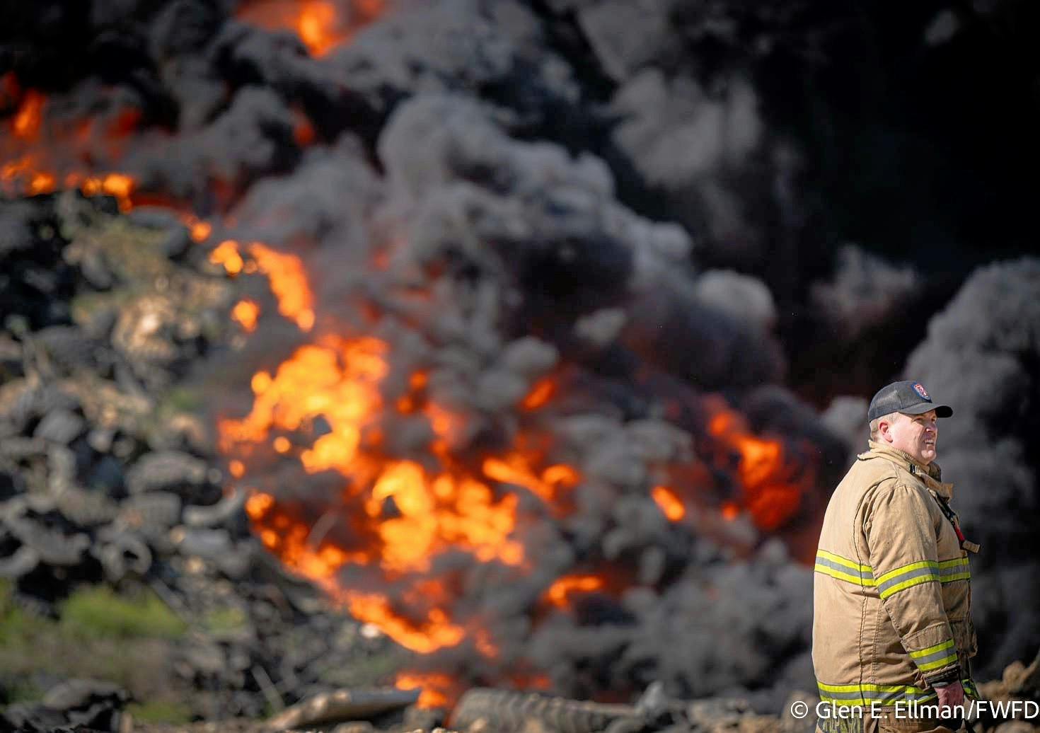 A Fort Worth firefighters watches over the scene as a large pile of tires and other debris burns in the background.